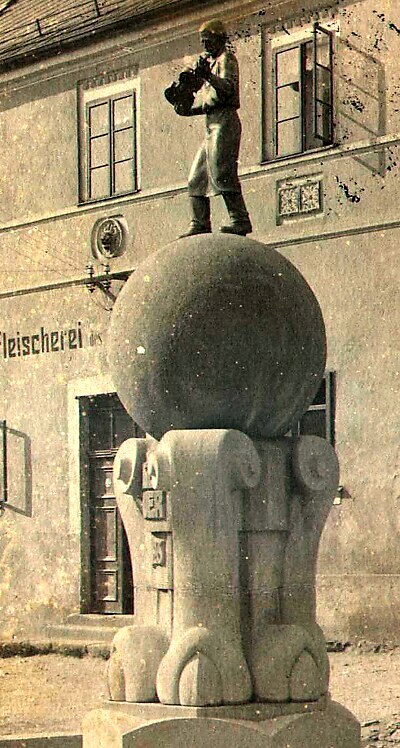 Sepia photograph of the violin makers’ monument: a standing figure holding a violin atop a large stone globe, in front of a pale building with “Fleischerei” lettering on the facade.