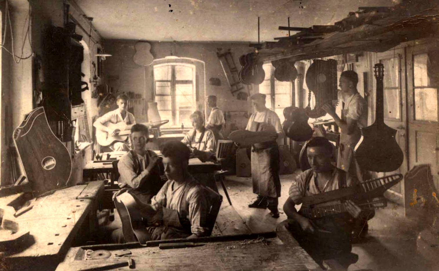 Sepia interior of a crowded early-20th-century instrument workshop: about eight people at wooden benches with guitars and tools, unfinished bodies hanging on walls and from ceiling beams, daylight from a rear window.