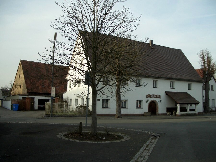 Colour photograph of a white two-storey traditional inn with a steep brown tiled roof on a street corner, bare winter trees, an arched doorway with a small porch, and an adjoining outbuilding with red roof tiles.
