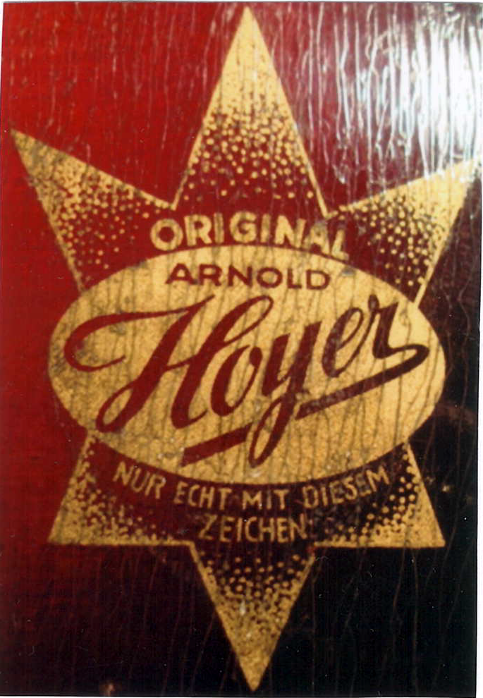 Close-up of a gold-stamped wooden authenticity label on reddish wood grain: a multi-pointed star around an oval reading “ORIGINAL ARNOLD Hoyer” and the German line “NUR ECHT MIT DIESEM ZEICHEN.”