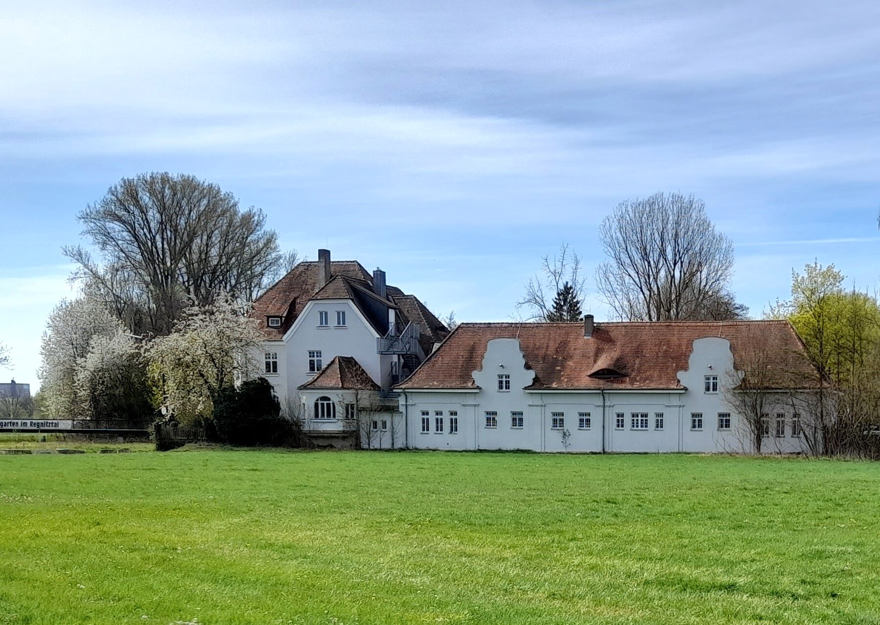 Colour photograph of a long white building complex with red-tiled roofs and shaped gables across a green meadow under a blue sky, with trees and spring blossom behind—the Erlangen Thalermühle.